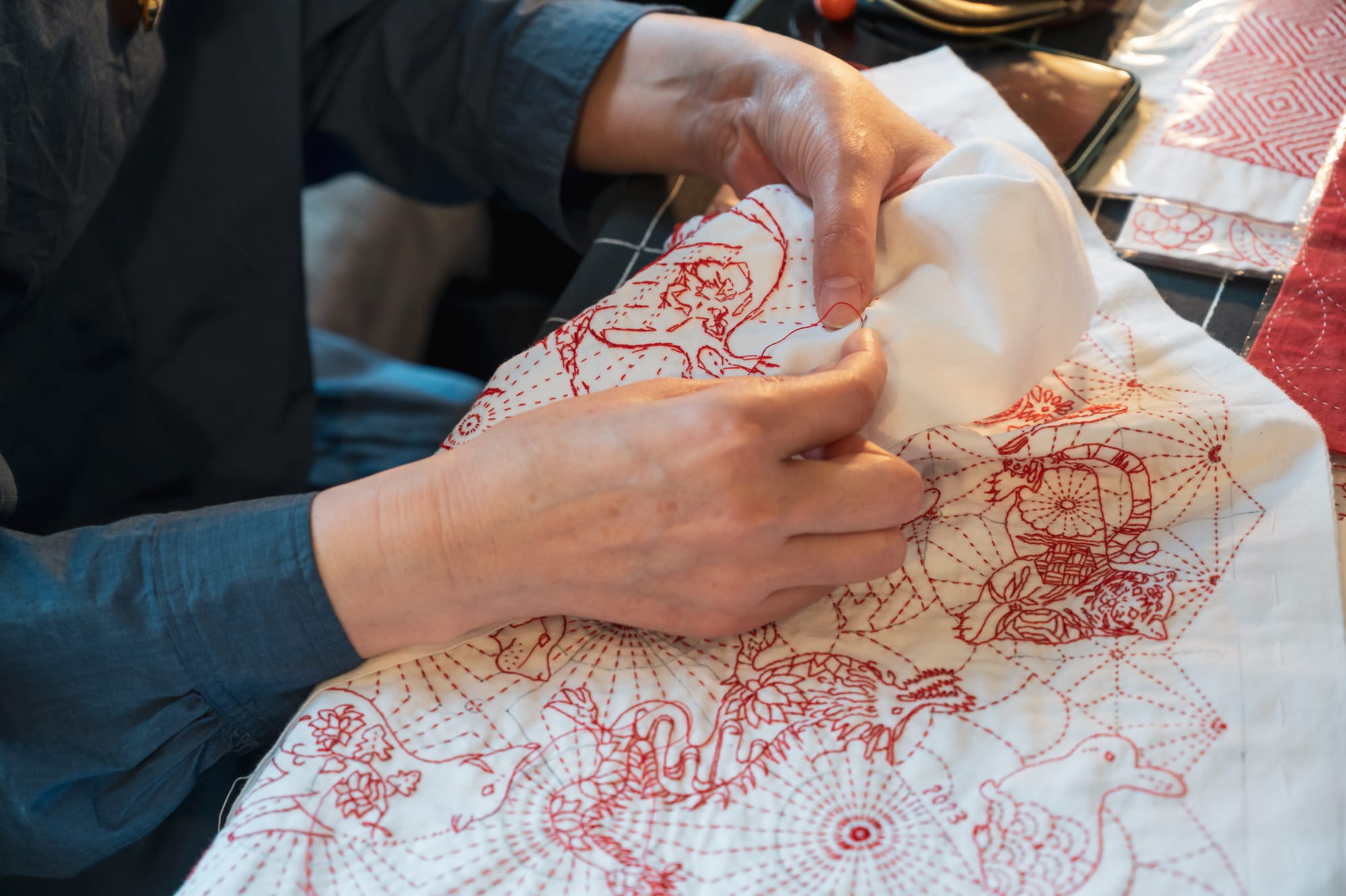 Chiebaa working on an intricate sashiko design showing several zodiac signs interwoven with traditional sashiko patterns in red on white